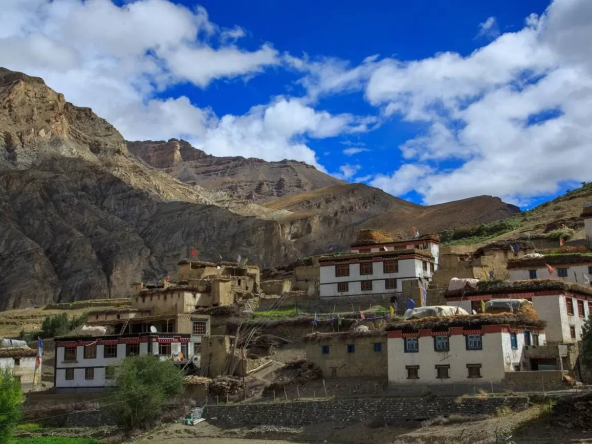 Hikkim Village Spiti near Kaza during partly cloudy skies, featuring white traditional houses rocky cliffs brown hills snow mountains, perfect cultural experience Himachal tour package.
