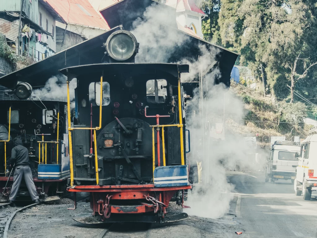Darjeeling Himalayan Railway steam engine at station during misty morning, featuring smoke clouds, hillside town and tracks, perfect Sikkim tour package