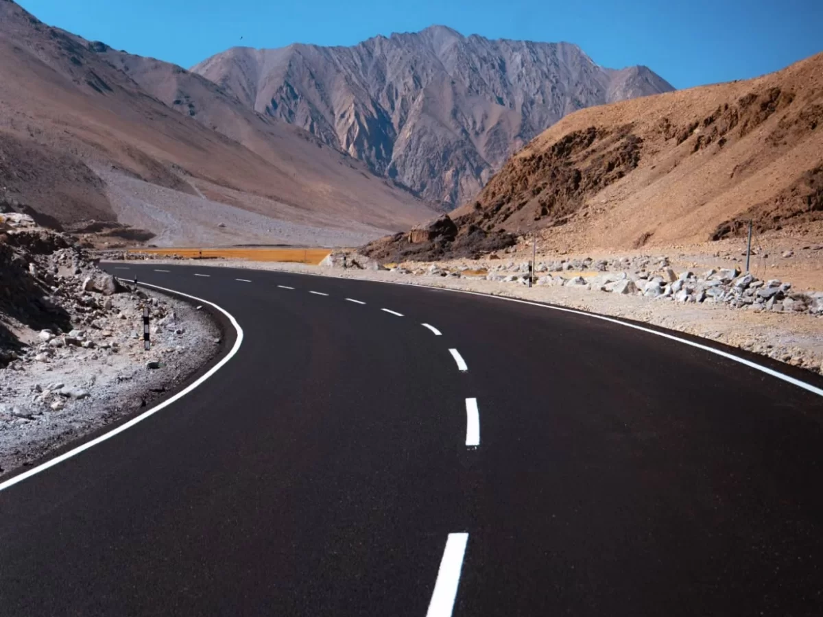 Winding road curves Hunder Nubra Valley Ladakh through dramatic barren mountains, blacktop highway with white center line snaking between rugged Karakoram peaks under clear blue skies, perfect adventure Ladakh tour package