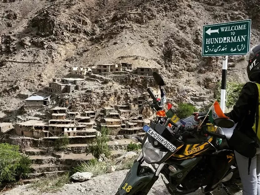 Hunderman Village Kargil Ladakh welcome signboard with motorcycle adventurer foreground, ghost village cliff houses on barren mountains, Brokpa settlement LoC border, perfect Himalayan adventure India tour package. 