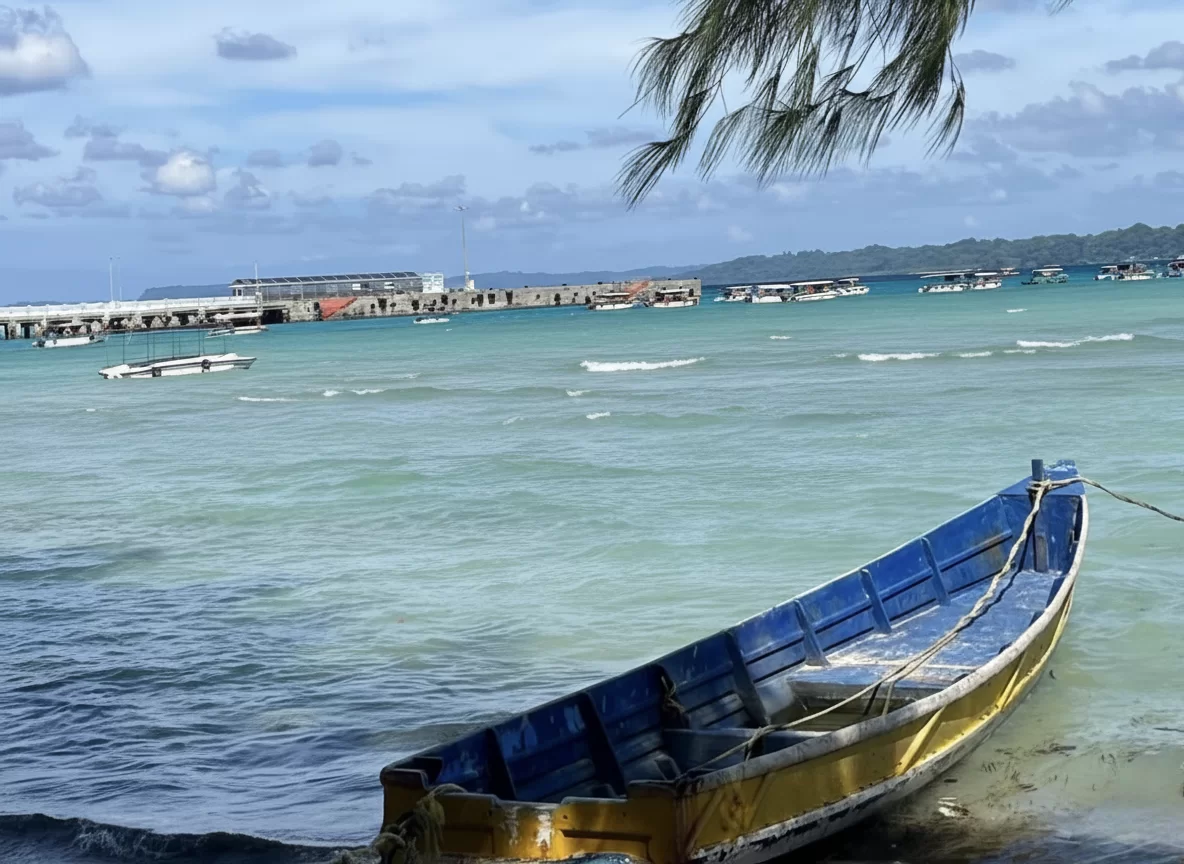 Bharatpur Beach Port Blair Andaman Nicobar traditional blue-gold fishing boat anchored in turquoise lagoon, palm trees, distant jetty with boats under partly cloudy tropical skies, perfect eco beach Andaman tour package.