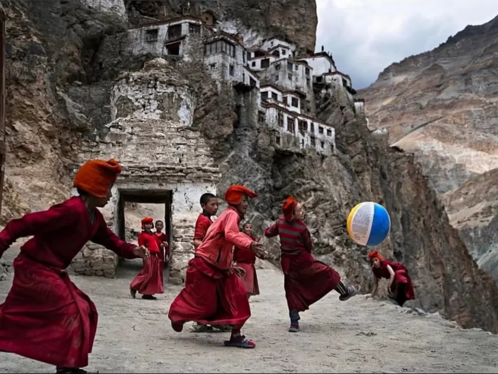 Ladakh Zanskar Phugtal Gompa cave monastery white building cliff face red robed Buddhist monks novices playing blue beach ball saffron hats traditional robes rocky barren mountain landscape cloudy sky remote pilgrimage adventure trek tourism photography. 