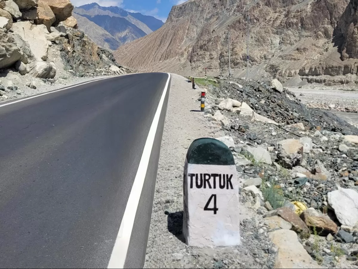 Road milestone to Turtuk village in Nubra Valley Ladakh during clear day, featuring Turtuk 4 km sign, rugged mountains, riverbed, perfect adventure Nubra tour package.
