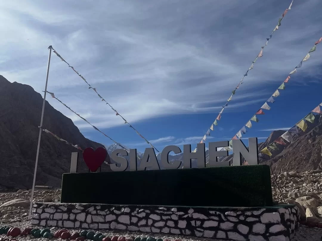 I Love Siachen large signboard red heart white letters prayer flags fluttering poles Karakoram mountains cloudy sky rocky terrain tourist photo spot Siachen Base Camp Ladakh Nubra Valley. (162 chars) ​
