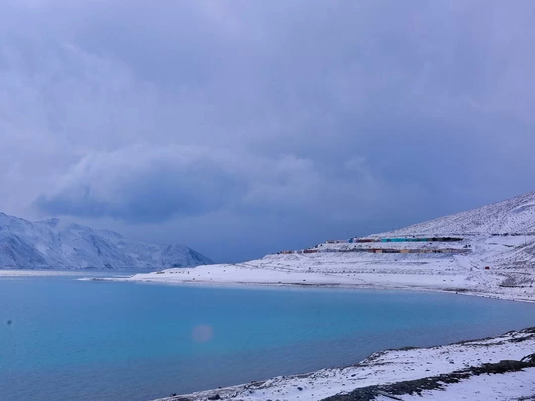 Pangong Tso Lake Ladakh winter snowy landscape turquoise water blue ice shore snow covered mountains terraced hills multi color train blue purple cloudy sky, high altitude frozen lake adventure tour package.