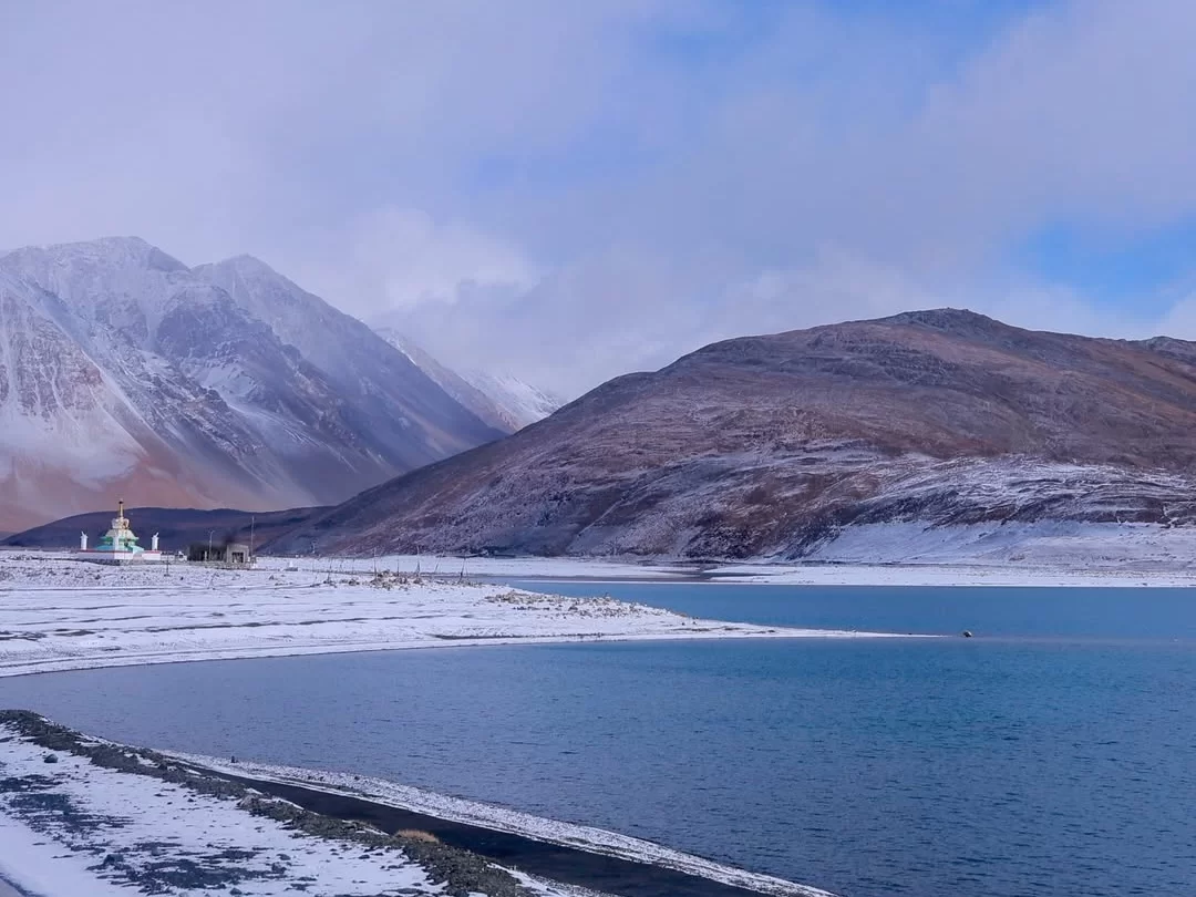 Pangong Tso Ladakh winter turquoise lake snow shore frozen edges green roofed monastery stupa snowy brown mountains blue purple cloudy sky distant peaks, high altitude Buddhist site winter tour package.