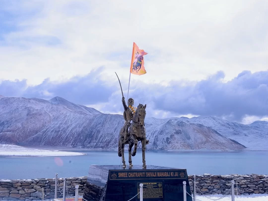 Pangong Lake Ladakh winter Chhatrapati Shivaji Maharaj statue bronze equestrian sword saffron flag pedestal snowy mountains turquoise water cloudy sky stone wall railing, historical monument adventure tour package. 
