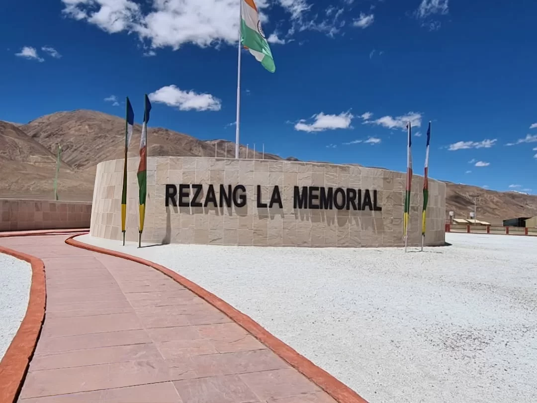 Rezang La War Memorial at Rezang La Pass Ladakh during clear skies, featuring Indian Tricolour flag, prayer flags, stone wall, Himalayan peaks, perfect patriotic experience with Ladakh tour package. 