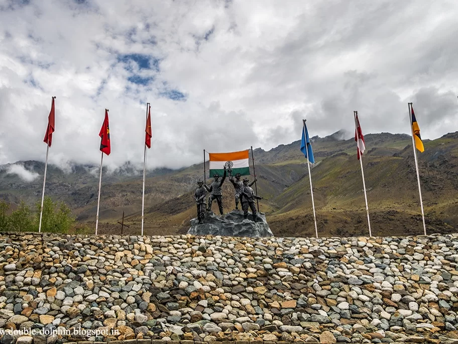 Drass War Memorial Ladakh soldiers statue Indian flags colorful banners snowy mountains rocky wall cloudy sky Kargil War heroes Operation Vijay tribute, perfect Ladakh heritage tour package.