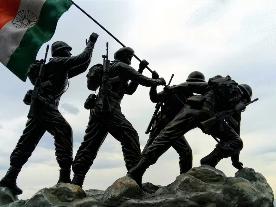 Drass War Memorial Ladakh soldiers statue Indian flag waving rifles helmets backpacks rocky base cloudy sky Kargil War heroes Operation Vijay tribute, perfect Ladakh heritage tour package.