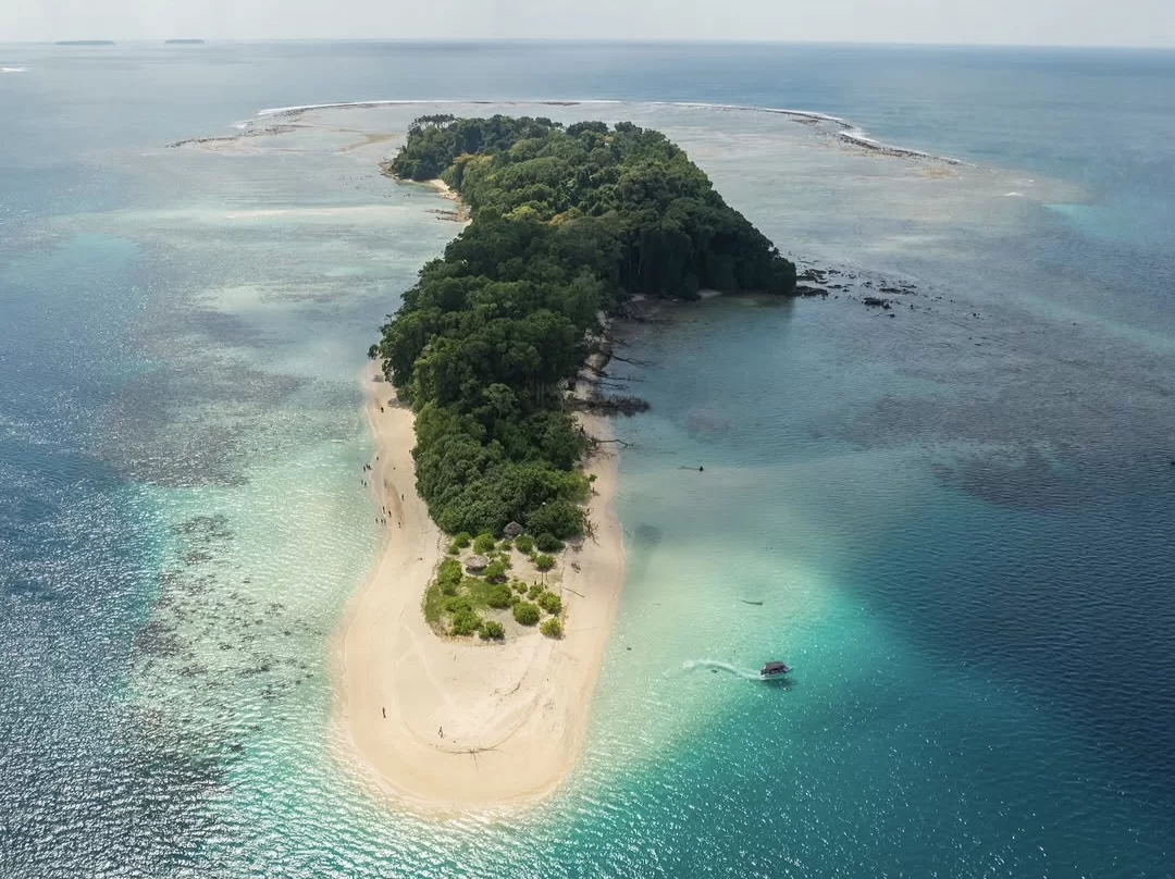 Aerial view of Jolly Buoy Island Andaman during sunny day, featuring lush tropical forest, white sandy beaches, turquoise coral reefs, motorboat, perfect eco-snorkeling adventure Andaman tour package