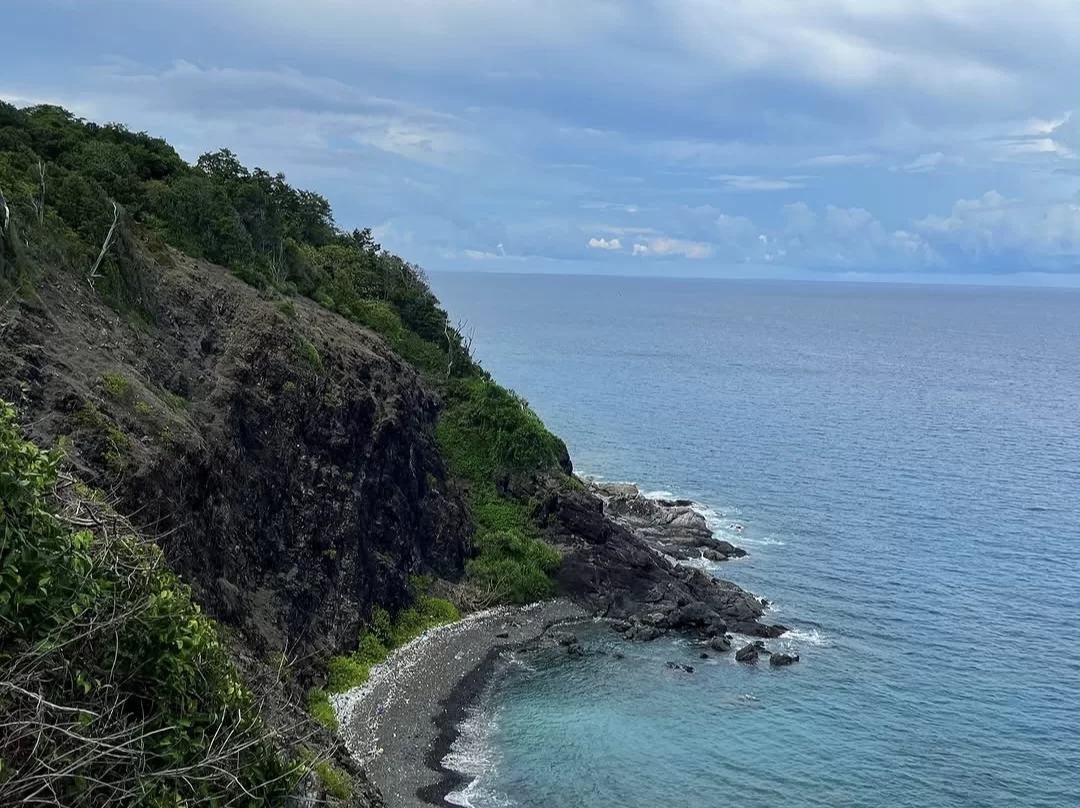 Cliffside view at Chidiya Tapu Port Blair during partly cloudy day, featuring black rocky cliffs, turquoise beach cove, lush green vegetation, ocean waves, perfect nature adventure Andaman tour package.