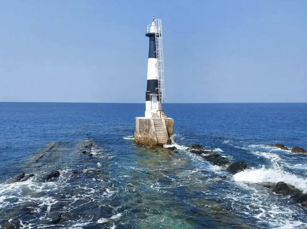 Black white striped lighthouse on rocky breakwater at Ross Island Andaman during clear partly cloudy day, featuring crashing turquoise waves, blue ocean horizon, perfect adventure Andaman tour package.