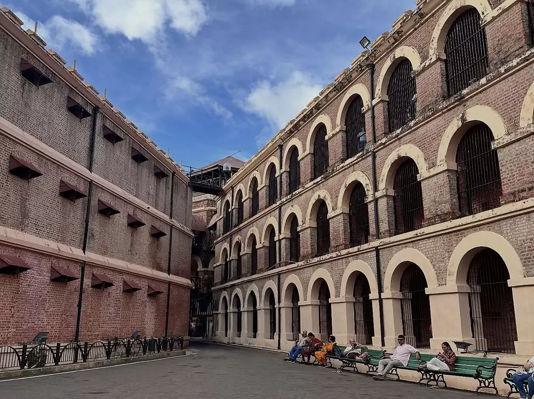 Cell blocks courtyard at Cellular Jail Port Blair Andaman during partly cloudy skies, featuring brick arches, iron grills, central watchtower, visitors, perfect heritage experience Andaman tour package.