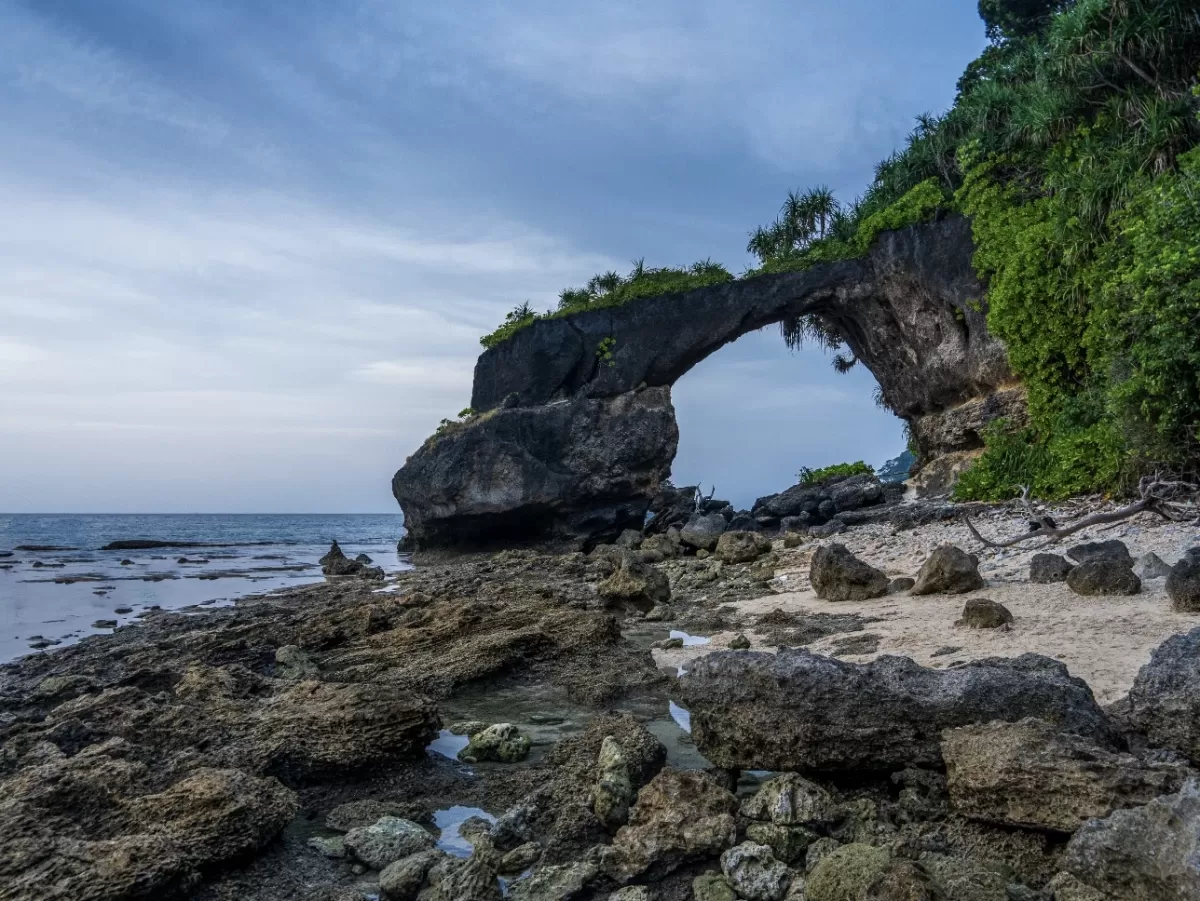 Natural limestone bridge rock formation amidst lush tropical vegetation rocky shore and calm sea at Laxmanpur Beach Neil Island Andaman under partly cloudy skies, signature natural arch adventure Andaman tour package feature