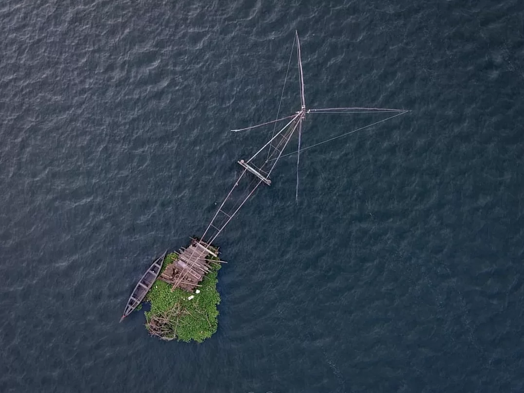 Aerial view of Chinese fishing net lowered into Vembanad Lake at Fort Kochi Kerala, featuring traditional cheena vala raft with trees, calm waters, perfect Kochi backwaters tour package. 