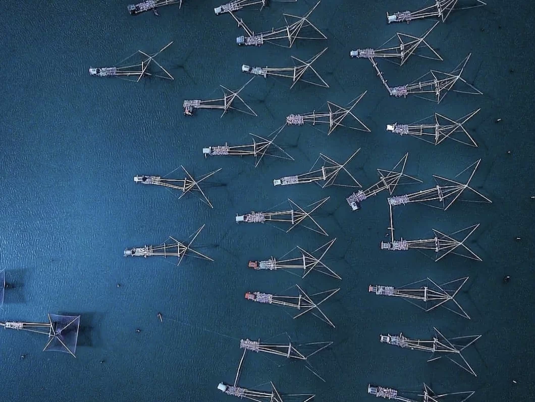 Aerial drone view of multiple Chinese fishing nets array over Vembanad Lake at Fort Kochi Kerala during twilight, featuring cantilever structures, deep blue waters, perfect Kochi tour package.