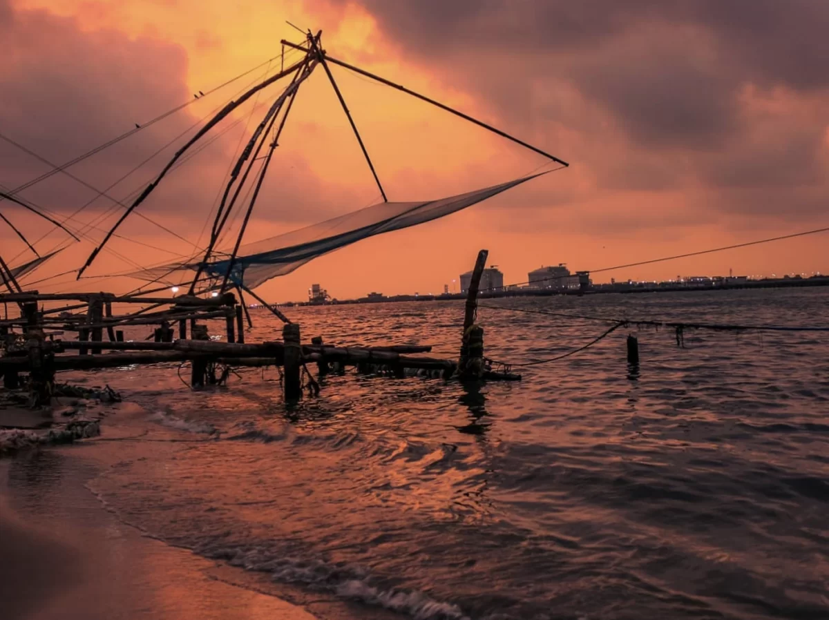 Chinese fishing nets silhouette at Fort Kochi Kerala during fiery orange sunset, featuring dramatic cloudy sky, cantilever nets over Vembanad Lake shore, perfect romantic Kochi tour package.