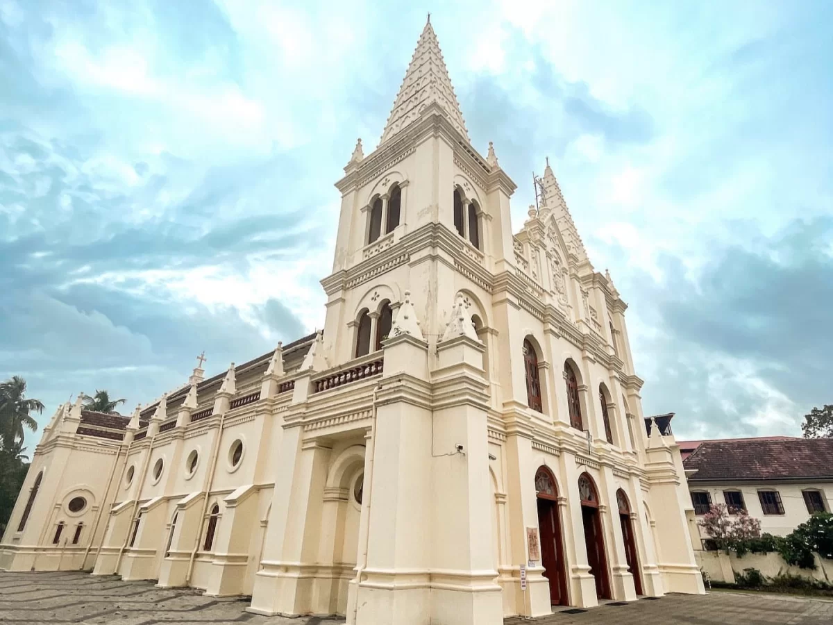 Kochi Kerala Santa Cruz Basilica Cathedral white Gothic architecture twin spires arched windows red doors balustrade cloudy sky palm trees traditional Kerala roof building heritage church tour package. 
