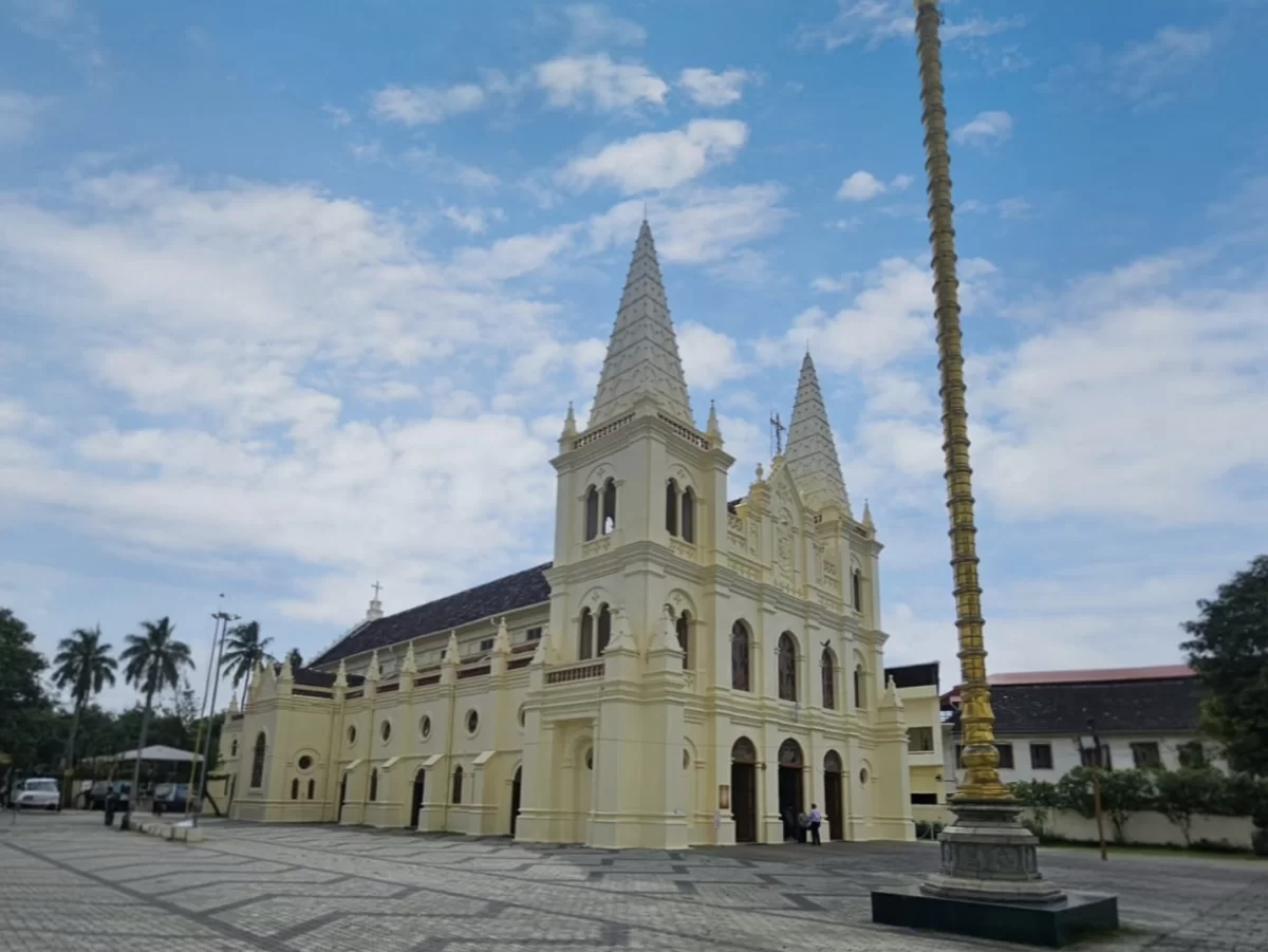 Kochi Kerala Santa Cruz Basilica Cathedral pale yellow Gothic twin spires arched windows golden flagstaff pole pedestal square plaza palm trees blue sky scattered clouds people Kerala roof building, colonial heritage church tour package.