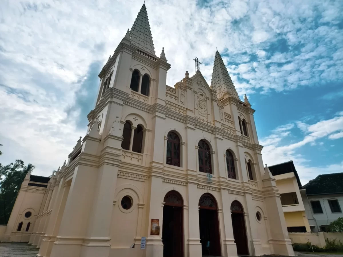 Kochi Kerala Santa Cruz Basilica Cathedral cream white Gothic Revival architecture tall pointed spires crosses arched windows brown doors trees cloudy blue sky traditional buildings foreground, historic Portuguese church heritage tour package. 