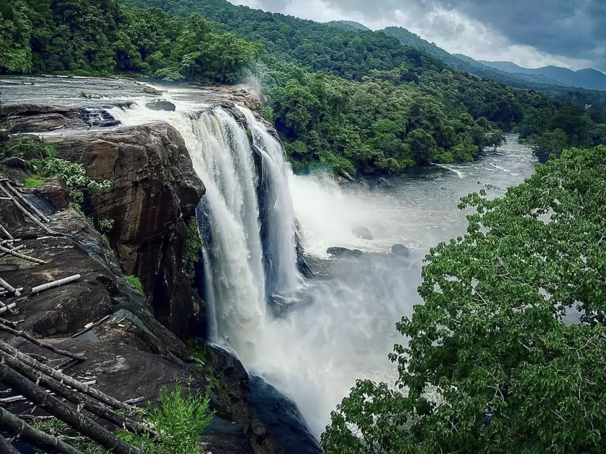 Athirappilly Waterfalls Thrissur Chalakudy Kerala India stunning wide cascading waterfall tumbling dark rocky cliff Chalakudy River dense green rainforest hills misty foam cloudy overcast skies foreground, perfect Kerala monsoon nature trekking tour packa