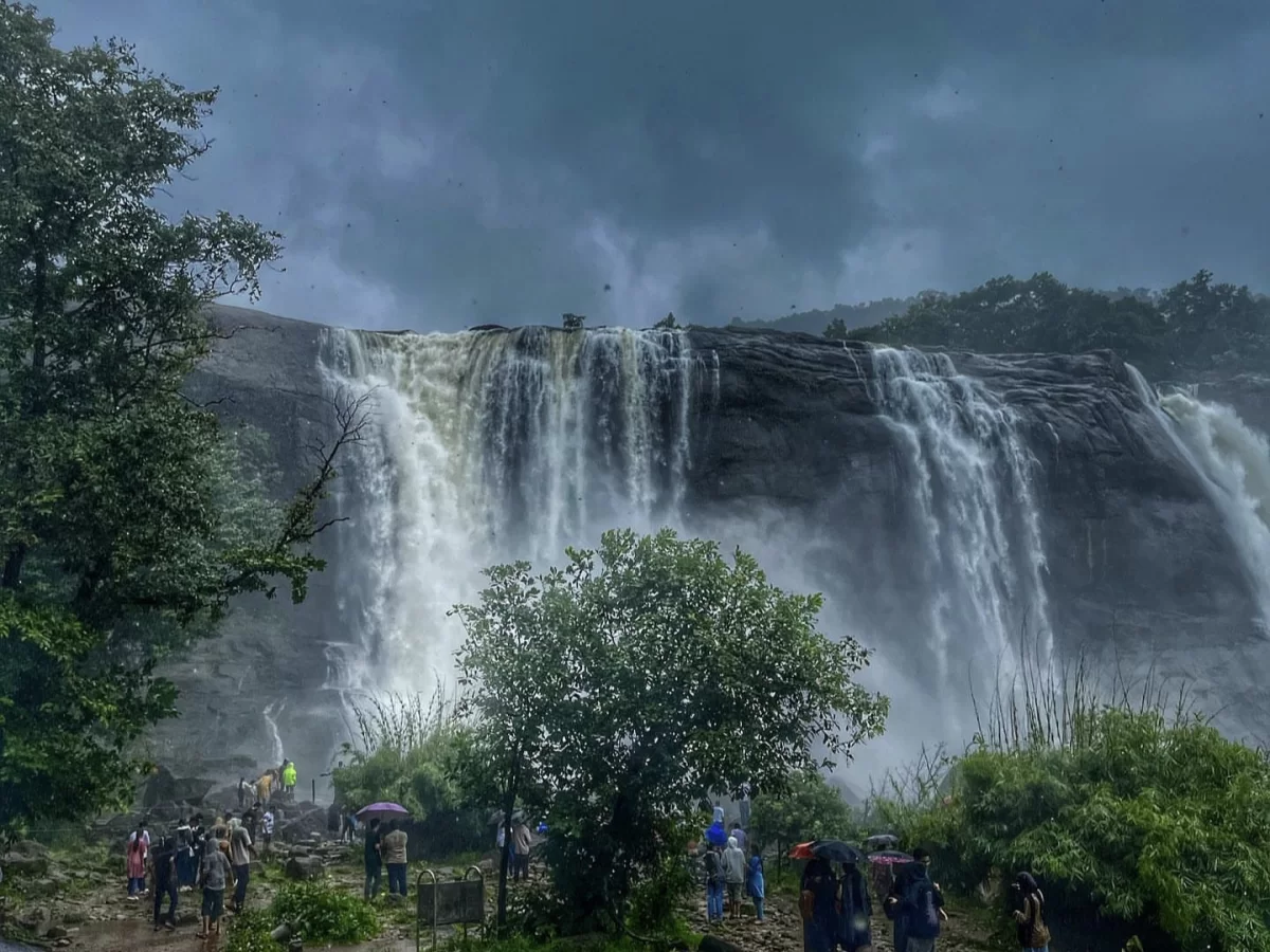 Athirappilly Waterfalls Thrissur Chalakudy Kerala India breathtaking multi tiered waterfall plunging massive rock face lush green trees rainy grey skies tourists umbrellas foreground misty spray vegetation, perfect Kerala waterfall monsoon adventure tour 