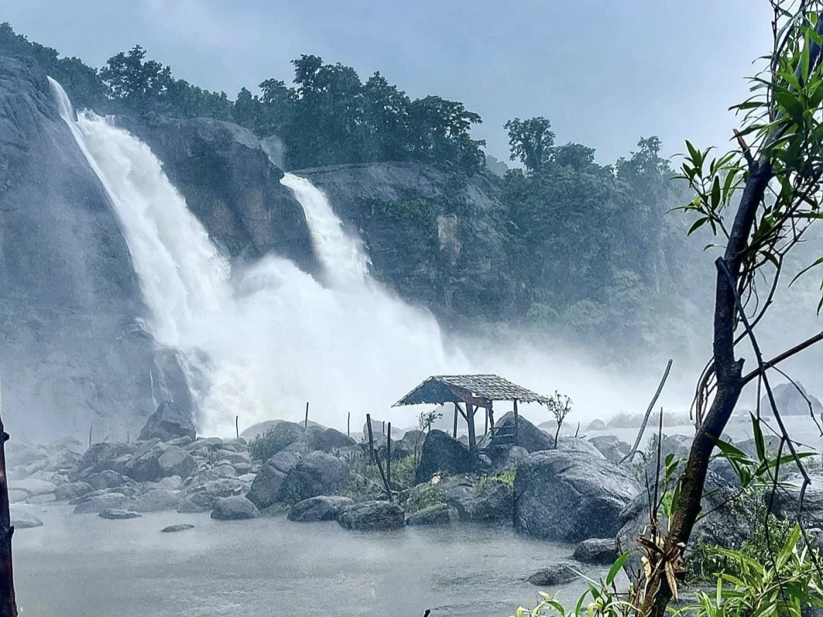 Athirappilly Waterfalls Thrissur Chalakudy Kerala India dramatic tall waterfall pouring rocky cliff lush rainforest overcast rainy skies foreground bamboo hut rocks shallow pools green foliage, perfect Kerala nature photography tour package.