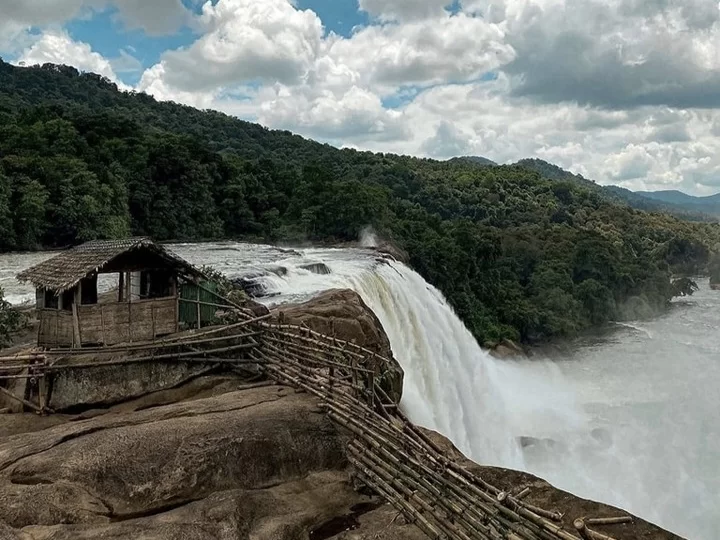 Athirappilly Waterfalls Thrissur Chalakudy Kerala India scenic viewpoint bamboo thatched hut railing overlooking powerful waterfall Chalakudy River lush forested hills misty spray partly cloudy blue skies rocky cliff edge, perfect Kerala eco adventure tou