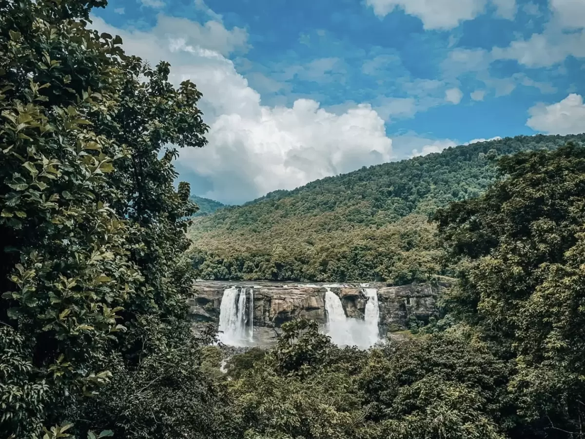 Athirappilly Waterfalls Thrissur Chalakudy Kerala India framed through lush green trees double tiered cascading waterfall rocky cliff Chalakudy River surrounded dense tropical forest mountains partly cloudy blue skies misty spray, perfect Kerala wildlife 