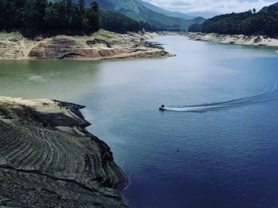 Munnar Kerala Mattupetty Dam reservoir green water speedboat wake brown terraced hills sandy shores lush evergreen forests distant misty mountains cloudy sky, hill station boating adventure tour package.