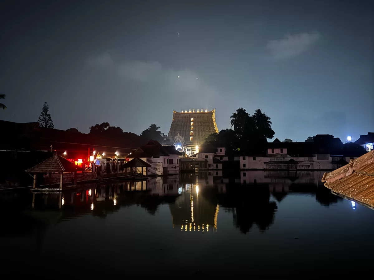 Sree Padmanabhaswamy Temple Thiruvananthapuram Kerala night illuminated golden seven-story gopuram lamps reflection temple tank water foreground palm trees huts red lights cloudy sky, Dravidian Hindu Vishnu shrine tour package. 