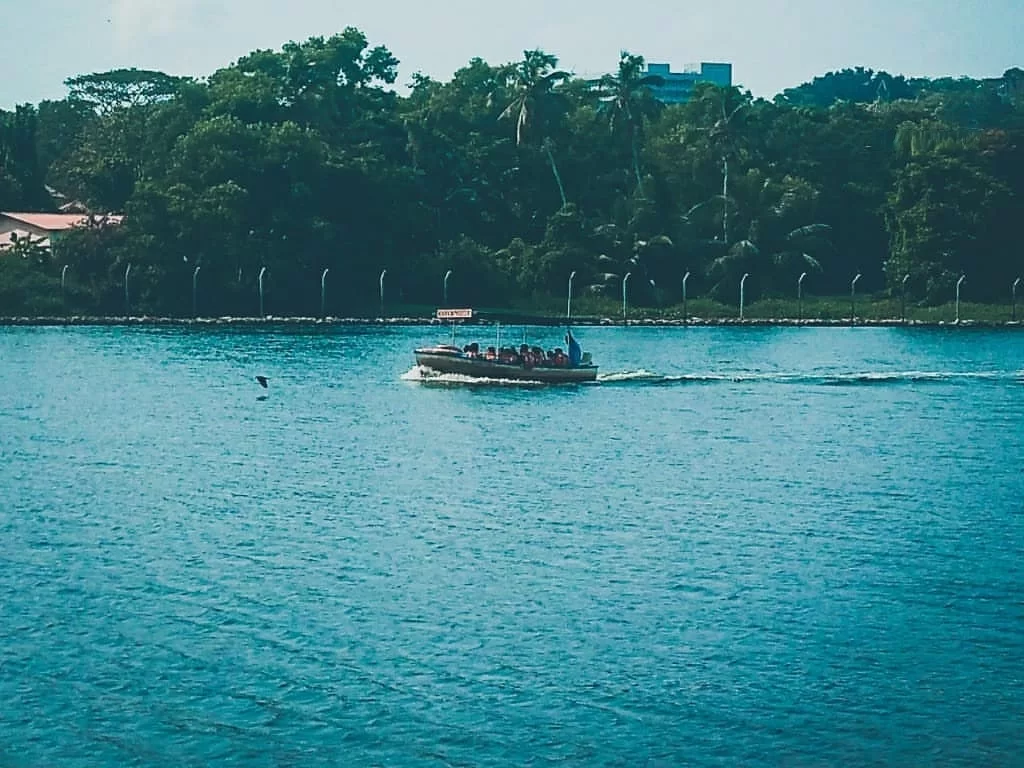 Thiruvananthapuram Kerala Veli Lake blue water tourist motorboat passengers palm trees green foliage shore red roof buildings distant city skyline serene backwater cruise tour package.