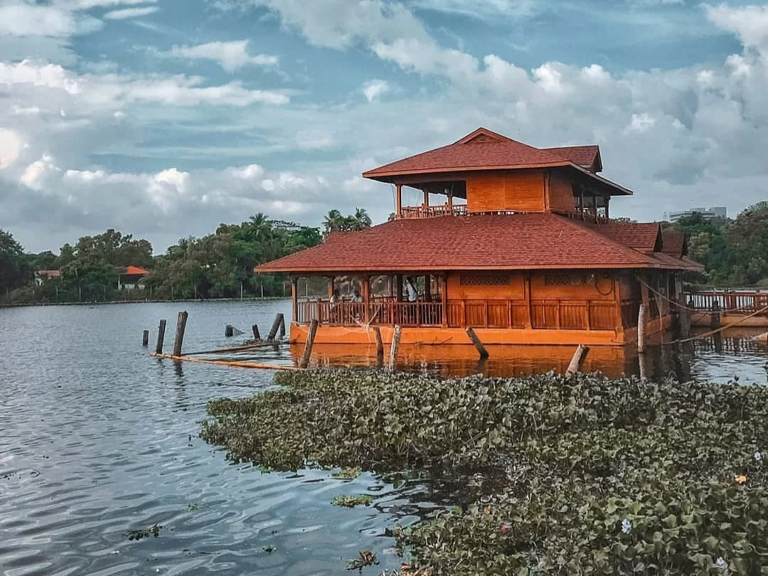 Thiruvananthapuram Kerala Veli Lake floating red Kerala style houseboat double tier balcony railings water hyacinth lily pads green trees cloudy blue sky serene backwater tourism tour package.