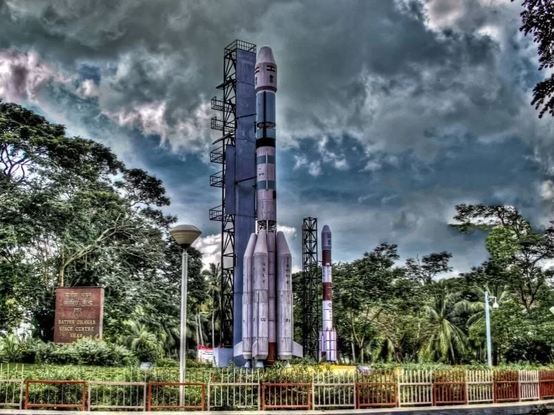 Thumba Space Centre Thiruvananthapuram Kerala ISRO rocket replica GSLV PSLV launch vehicle models gantry structure palm trees cloudy sky signboard fence lamp post, space museum science tour package.