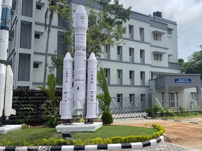 Thiruvananthapuram Kerala ISRO Space Centre entrance four white rocket models GSLV PSLV tricolor flag India Pro Hindi text grey building palm trees green fence lawn landscaping cloudy sky, space museum educational tour package