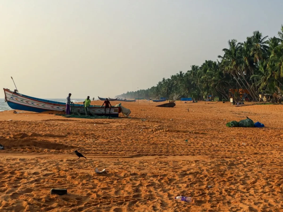Thiruvananthapuram Kerala Thumba Beach golden sand traditional blue painted fishing boats nets fishermen beach huts coconut palm trees hazy sunset sky serene coastal village scene tour package. 