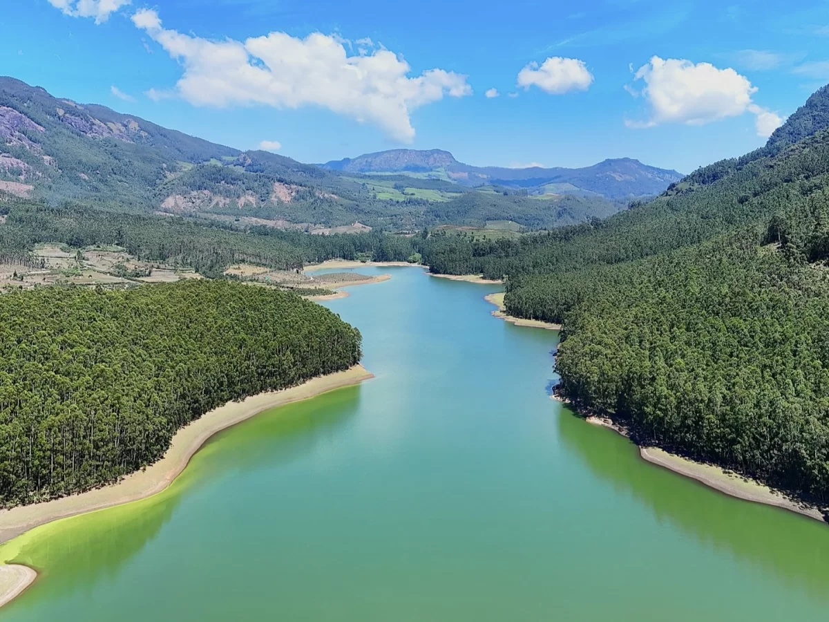Drone aerial view of Mattupetty Dam reservoir near Echo Point Munnar Kerala during sunny day with clouds, featuring turquoise green waters forested hills mountains yellow sandy shores lush eucalyptus trees, perfect panoramic nature experience with Munnar 