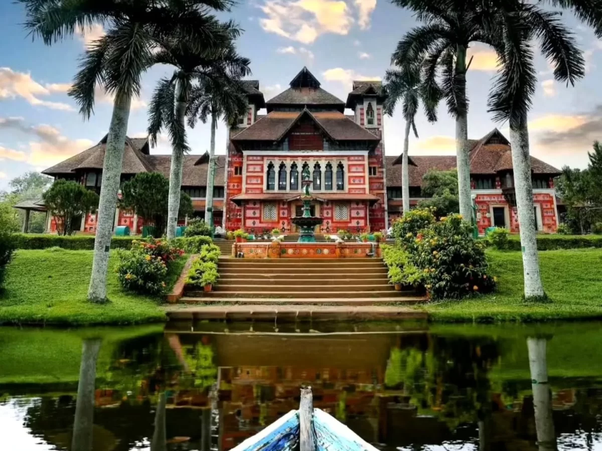 Napier Museum Kerala-style red-white palace with tall towers, arched verandas and central fountain reflected in pond, framed by coconut palms and gardens at dusk with boat foreground, perfect India tour package. 