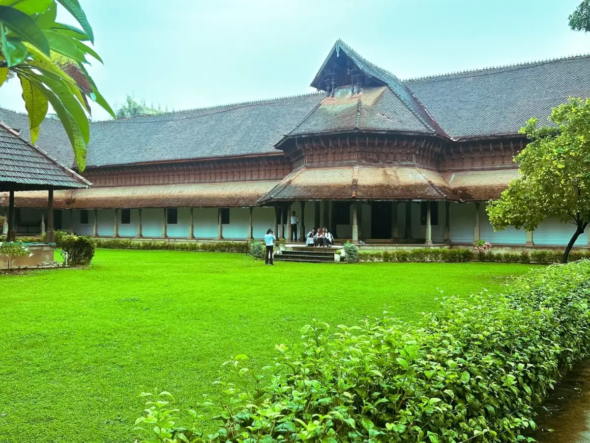 Kuthiramalika Palace Thiruvananthapuram Kerala courtyard scene with terracotta roofs, wooden pillars, visitors on steps amid lush green lawns under overcast sky, perfect India tour package.