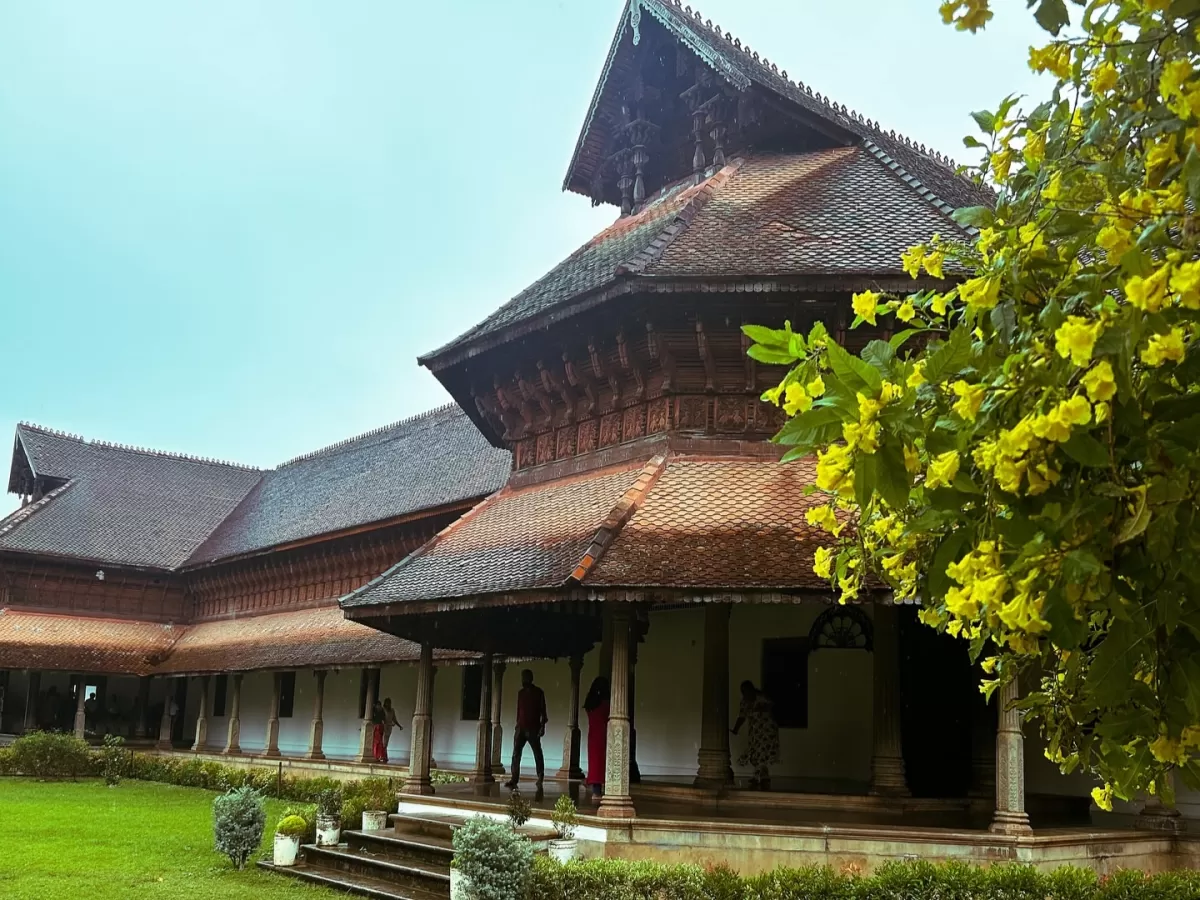 Kuthiramalika Palace Thiruvananthapuram Kerala ornate gopuram-style tower, terracotta roofs, yellow champa flowers, visitors on veranda amid green lawns under cloudy sky, perfect India tour package.