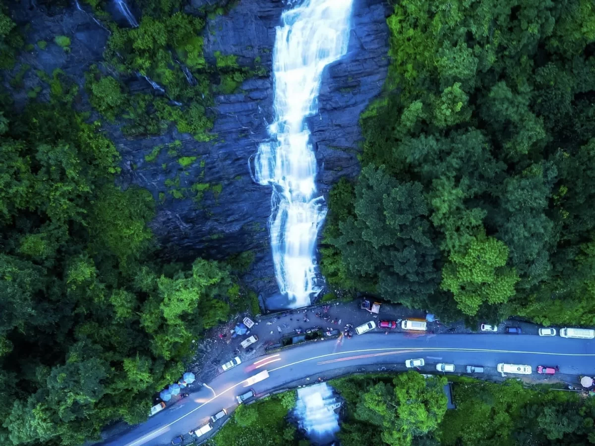 Aerial drone view Valara Waterfalls Kerala lush rainforest, featuring towering cascade winding road tourist vehicles dense greenery, perfect Kerala waterfall adventure tour package.