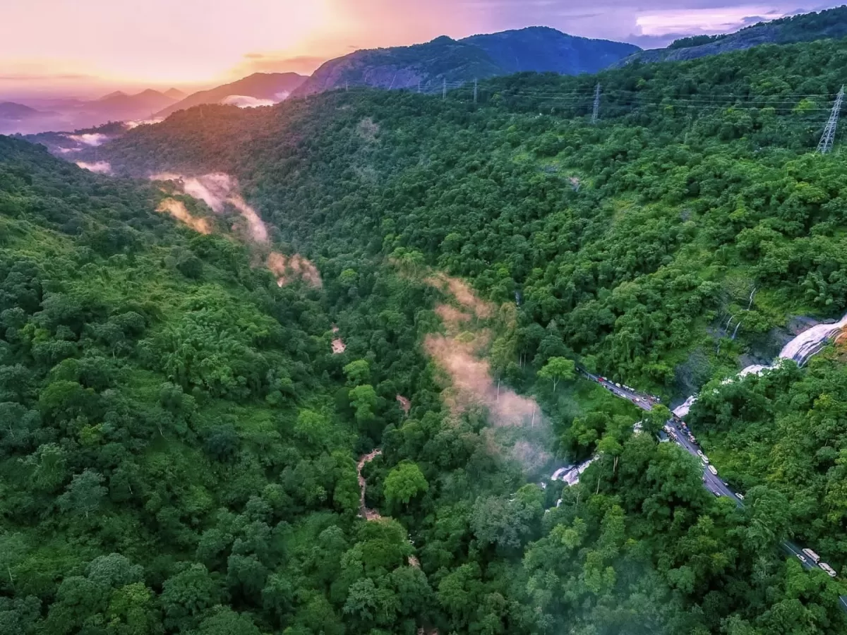 Drone aerial golden hour Valara Waterfalls Kerala valley, featuring misty cascades lush green forests winding roads power lines distant mountains, perfect Kerala nature tour package.