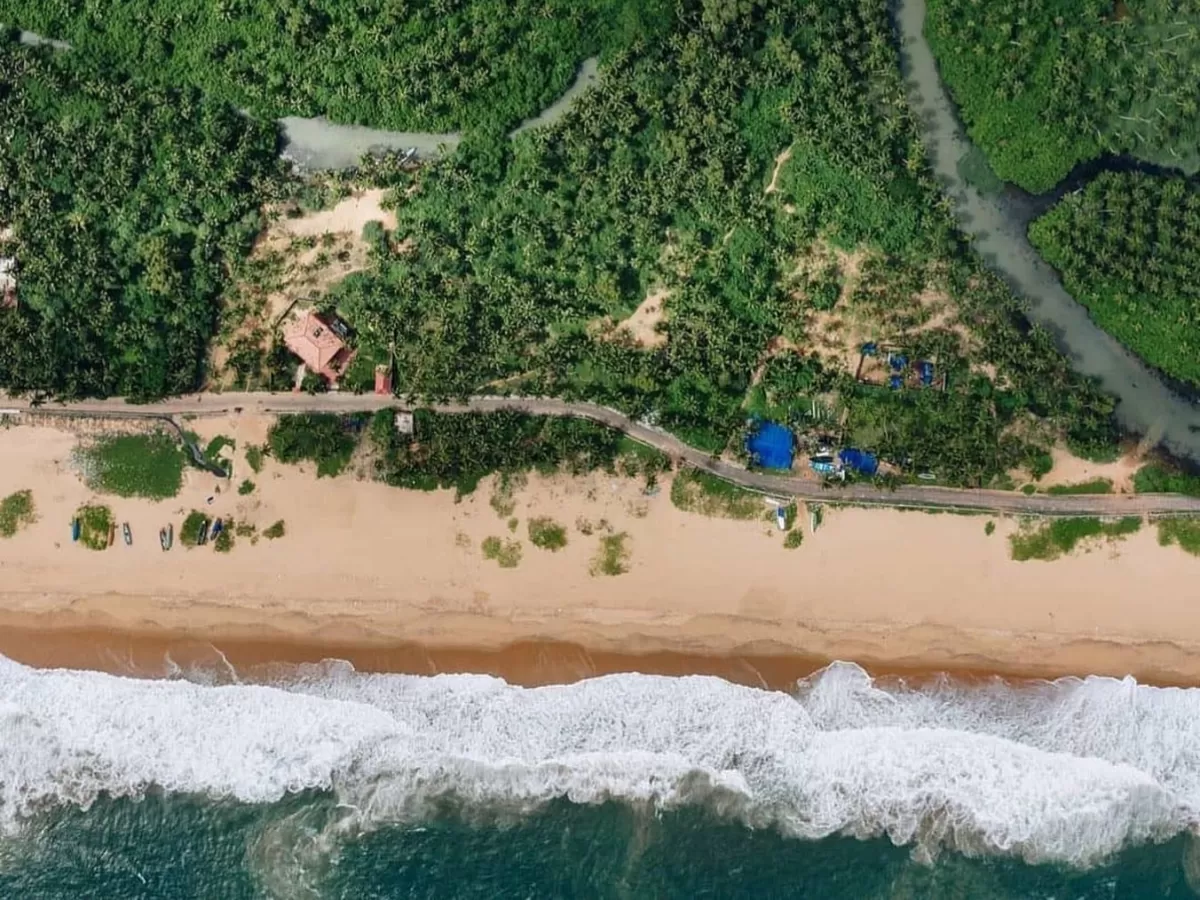 Aerial view of Poovar Island Kerala golden sandy beach meeting lush green coconut groves and mangrove backwaters with pink resort building blue pools houseboats road waves crashing into Arabian Sea, perfect India Kerala beach resort tour package. 