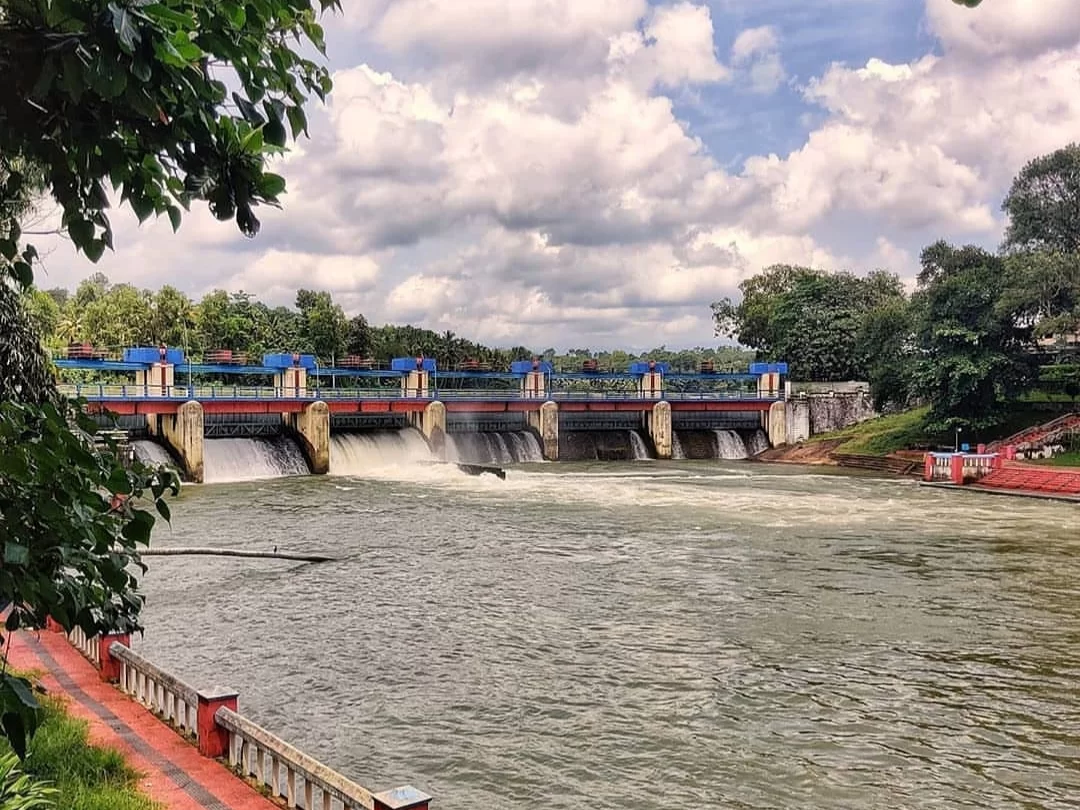 Aruvikkara Dam with water spilling over during partly cloudy sky, featuring blue gates, trees, river, perfect Kerala tour package near Trivandrum.