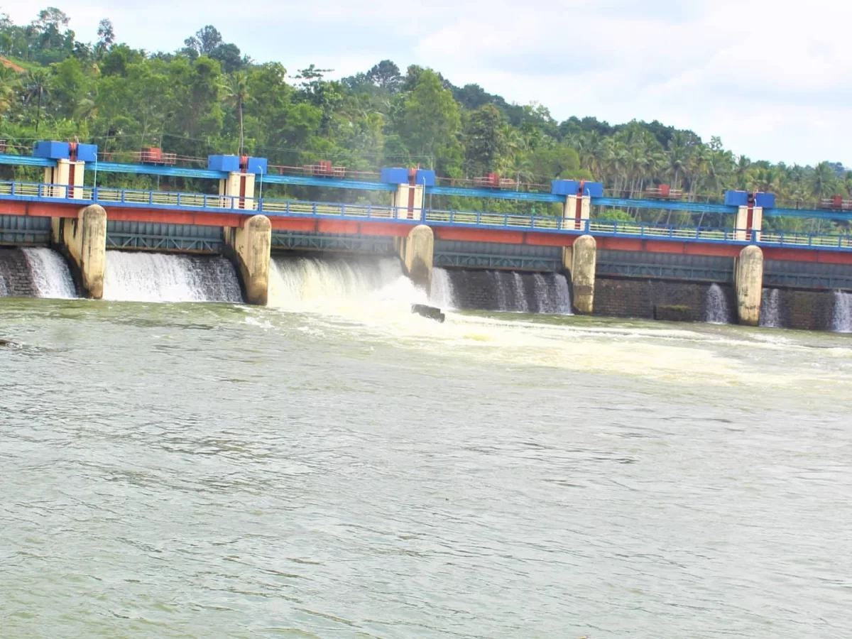 Aruvikkara Dam spillway with water release under blue gates amid lush trees and cloudy sky, perfect Kerala tour package near Trivandrum.