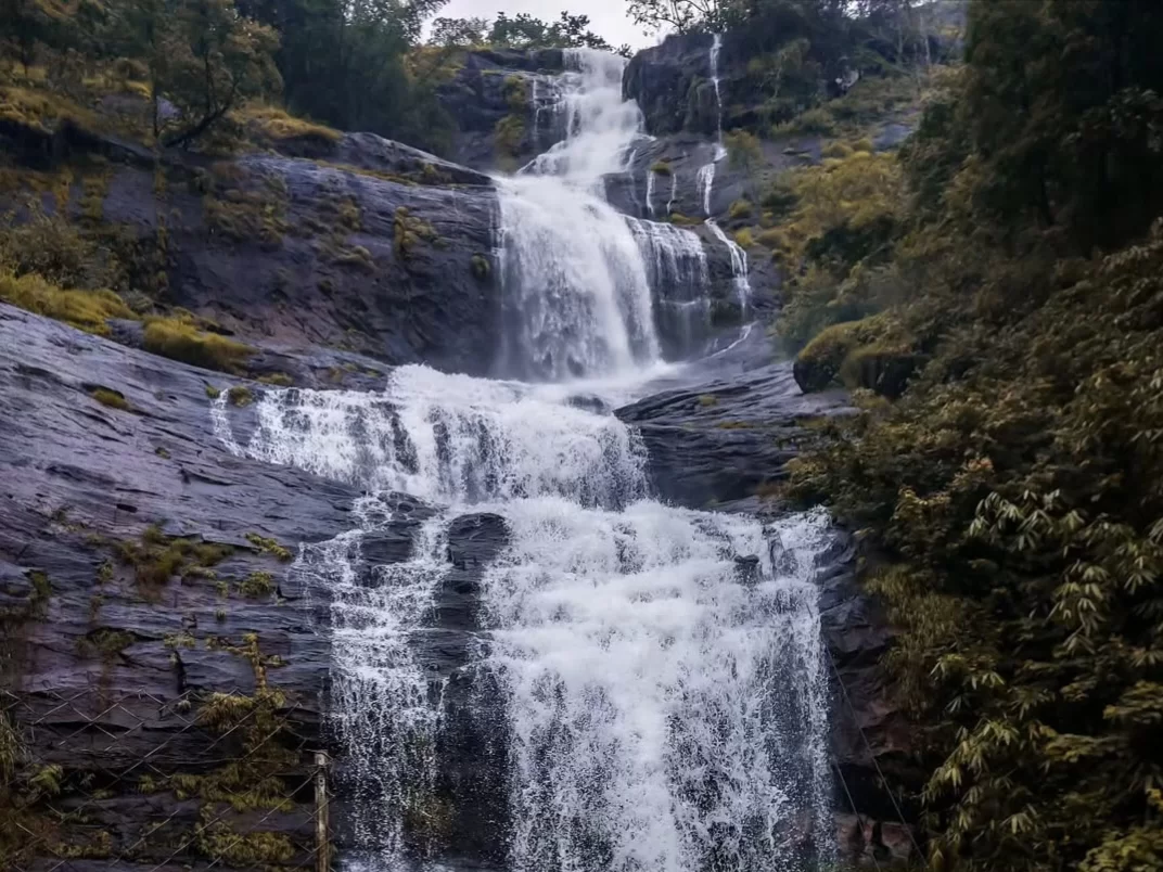 Multi-tiered Cheyyappara Waterfall near Munnar Kerala during overcast monsoon day, featuring gushing white cascades over dark rocky cliffs lush green forest, perfect roadside nature stop experience with Cheyyappara Waterfall Kerala tour package.