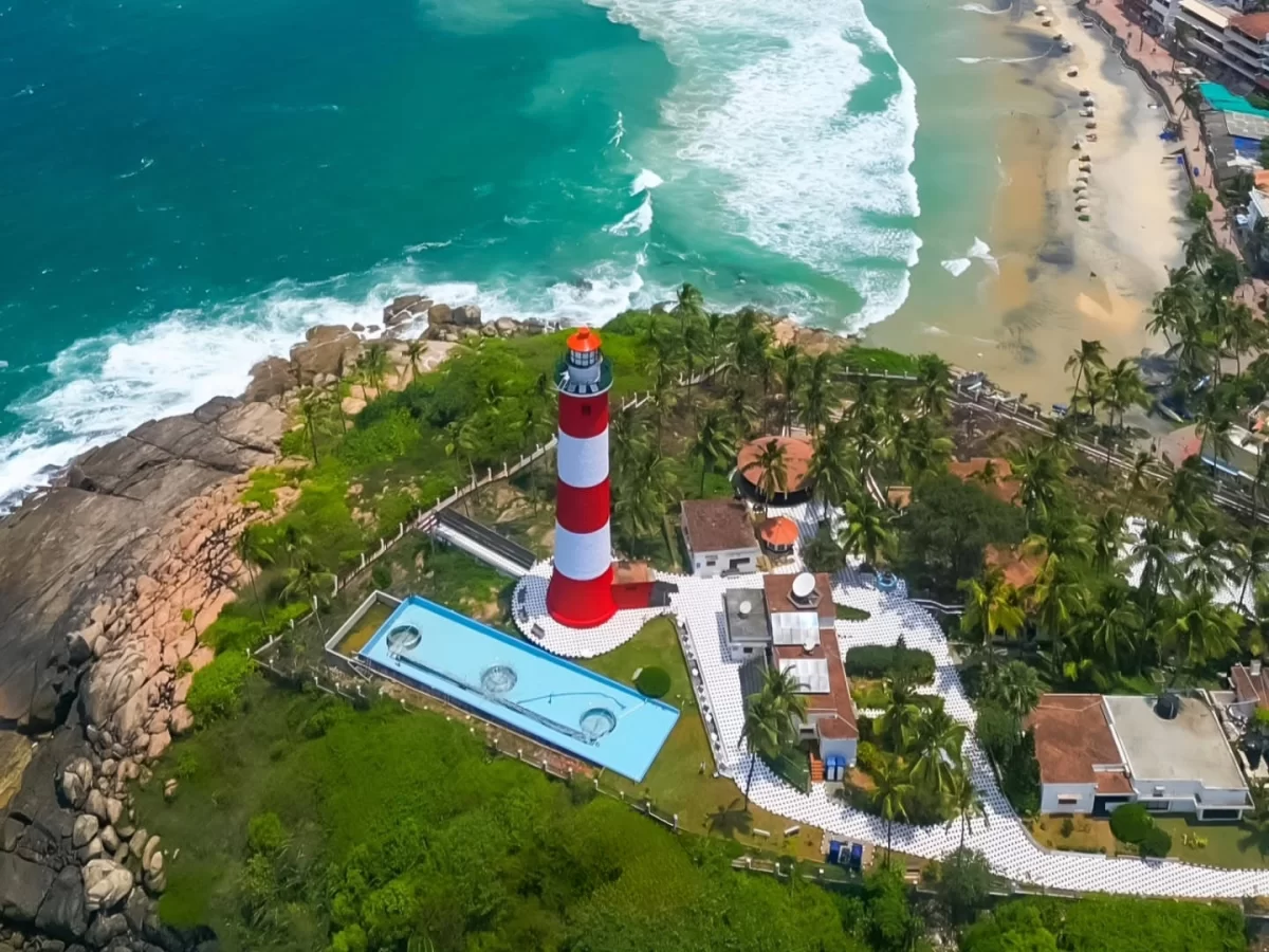 Aerial view of iconic red white striped lighthouse at Kovalam Beach Kerala perched on rocky promontory overlooking turquoise Arabian Sea waves, golden sand, coconut palms and infinity pool, perfect India tour package.