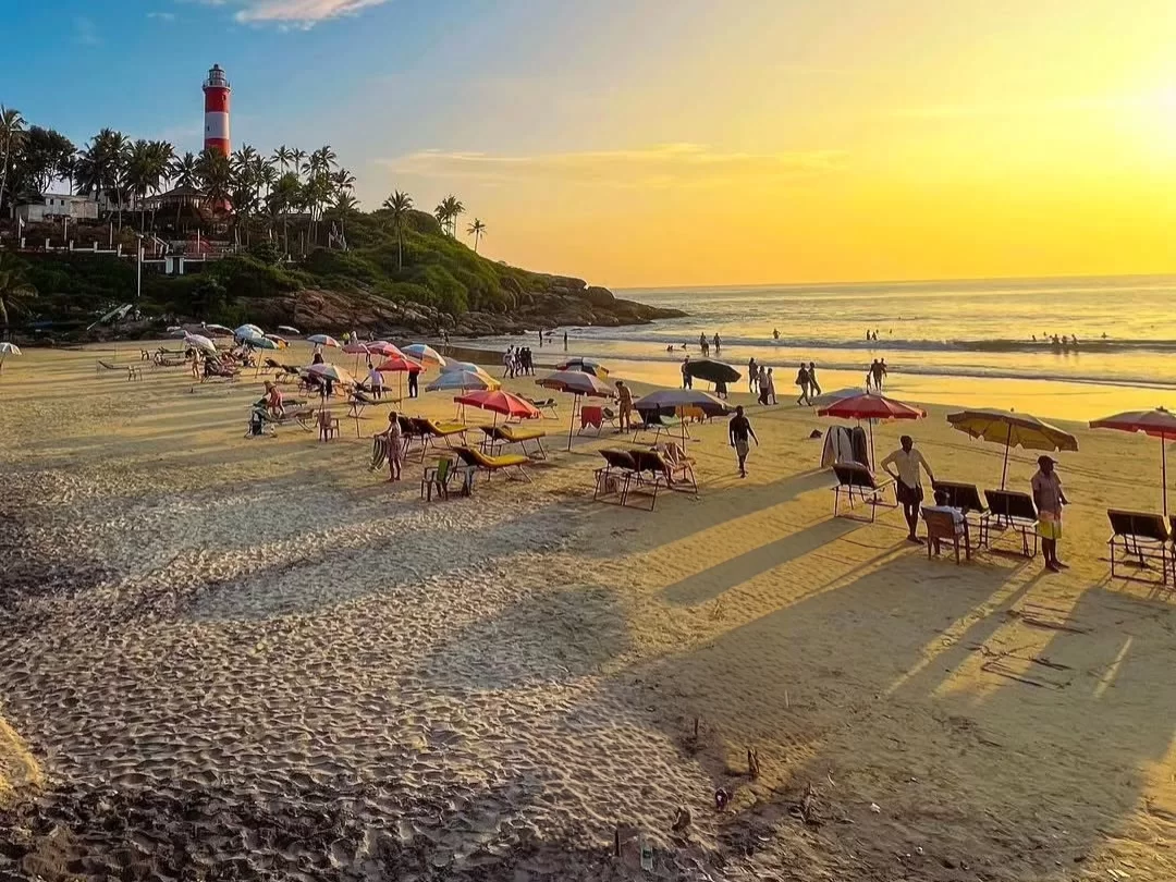 Golden hour at Kovalam Beach Kerala with iconic red-white lighthouse on cliff, colorful beach umbrellas, loungers, tourists strolling golden sand and Arabian Sea waves, perfect India tour package. 