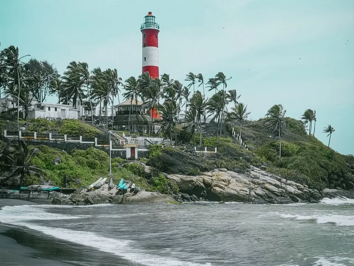 Panoramic view of Kovalam Beach lighthouse Kerala surrounded by swaying coconut palms, whitewashed buildings on lush green cliff overlooking dark Arabian Sea waves and sandy shore, perfect India tour package. 